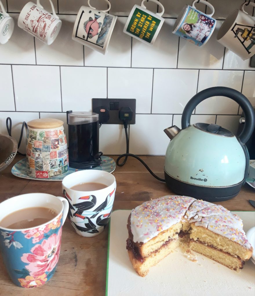 A wooden kitchen surface with a blue kettle, two filled cups of tea & victoria cake sponge on a white cake plate decorated with white icing and colourful hundreds & thousands sugar sprinkles. There are a row of colourful mugs hanging on the wll above a white tiled wall.