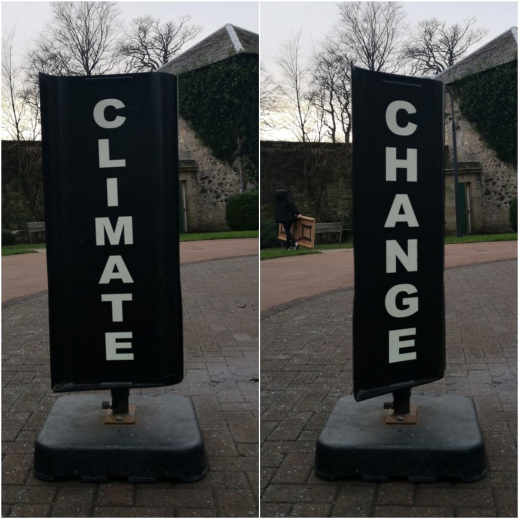 Two images collaged together side-by-side in one frame. The image on the left shows one side of a spinning black sign - the kind you would find outside a bureau de change. The sign vertically reads 'CLIMATE' in white capital letters. In the background, part of a building is visible against a pale blue twilight sky, which shows the silhouettes of leafless trees. The image on the right has exactly the same background, save for a person on the left walking away whilst carrying a wooden table. This time we see the other side of the sign, which is also black and vertically reads: 'CHANGE' in white capital letters.