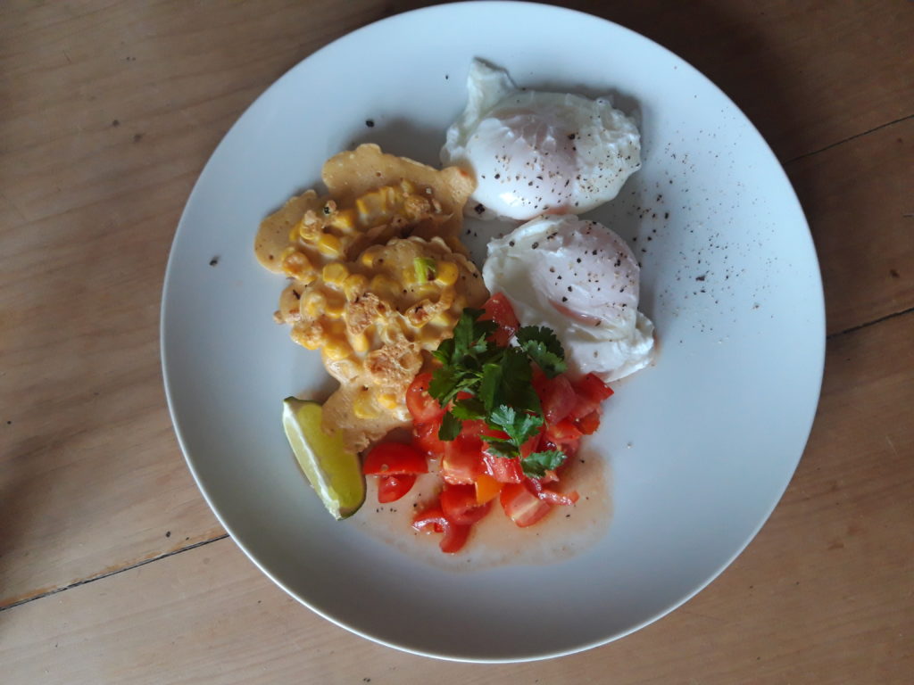 A plate of food. Fritters made of sweetcorn served with two poached eggs and tomato salsa on the side, garnished with coriander and a slice of lime.