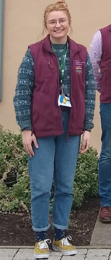 image shows a young woman, Kennedy Dold, standing smiling outside the Botanic Cottage. Kennedy is wearing the maroon volunteer gilet and has on the lanyard with keys for the Cottage. She is wearing jeans, a blue jumper, has blonde hair tied back and black rimmed glasses. 