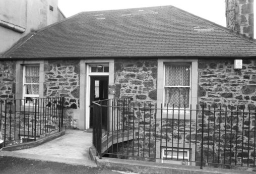 image shows a black & white image from the 1990's of the upper floor of the stone Cottage at road level with a front door and a window either side. You can just see a glimpse of the lower floor windows. There are black wrought iron railings at the front either side of the entrance way and the Cottage has a grey slate roof.