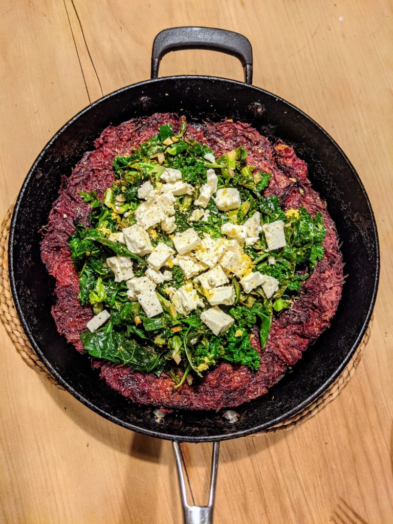 A pine table with a cast iron pan holding a Beetroot rösti with lemony greens and feta