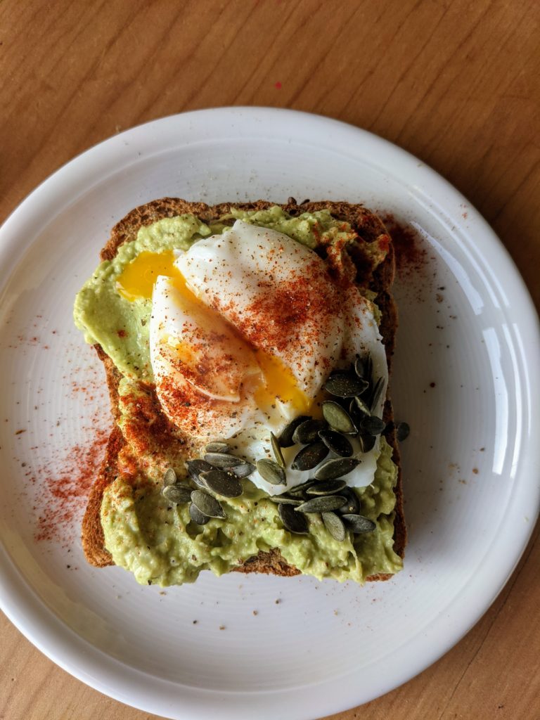 A white plate with a slice of toast topped with bright green avacadoa, a split poached egg garnished with smoked paprika