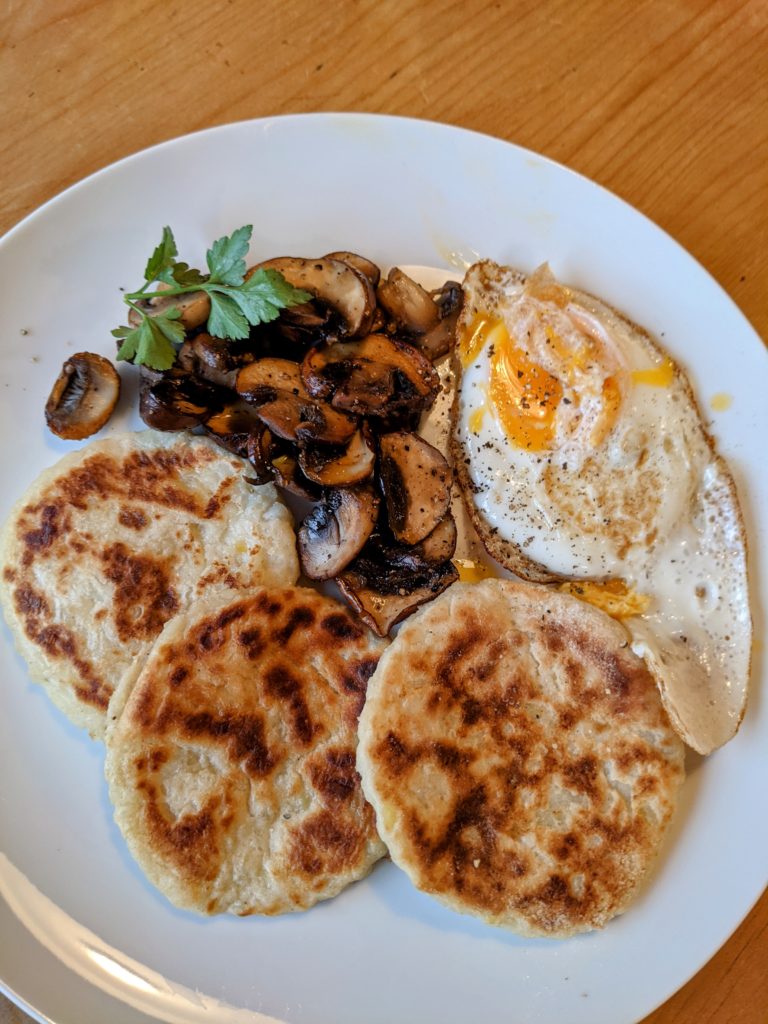 A white dish set with some round tattie scones fried eggs and fried mushrooms.