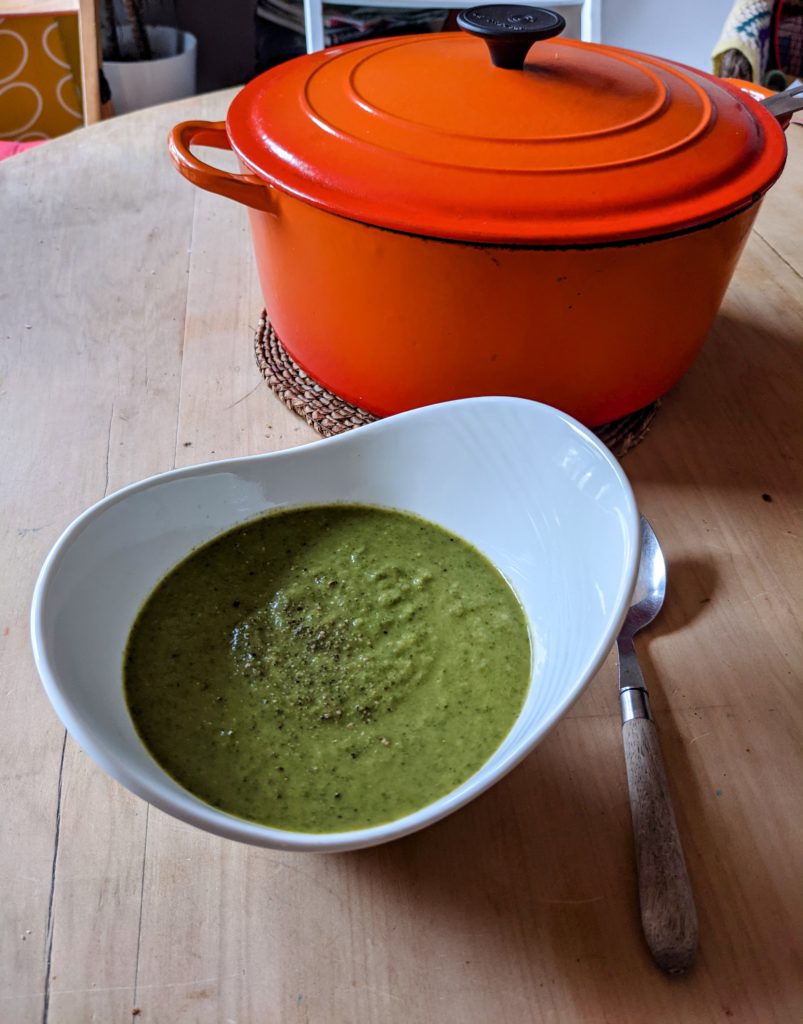 Bright red, large cooking pot on a wooden table with a white bowl of green courgette soup and spoon.