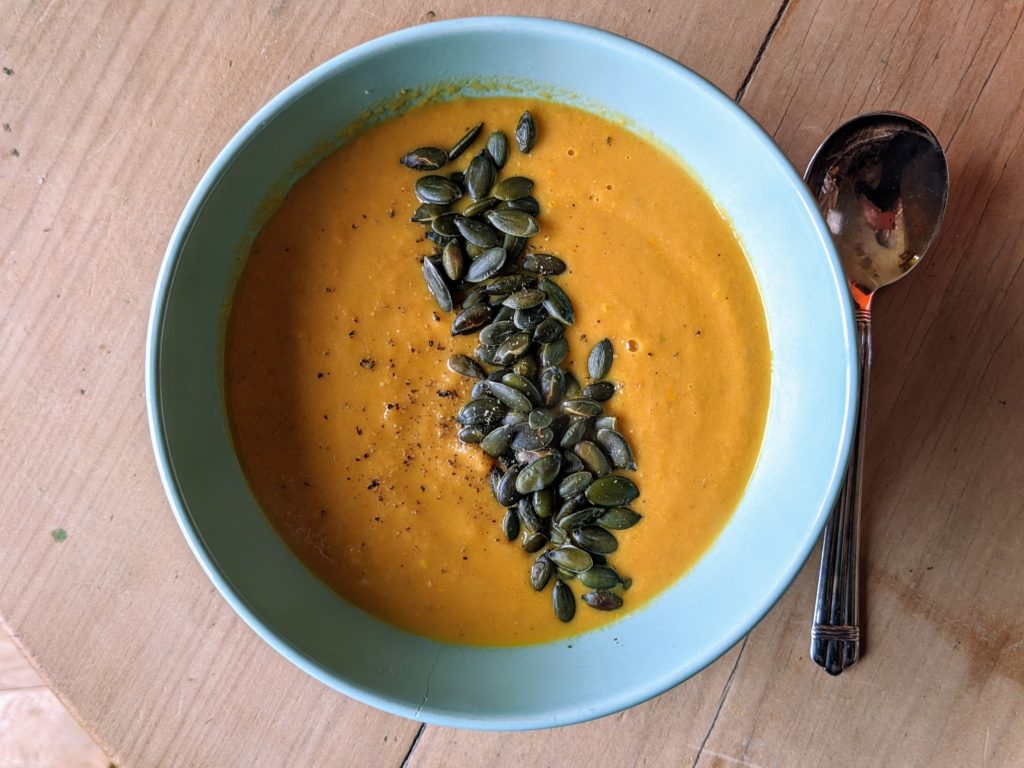 A white bowl on a pine table and silver spoon serving Carrot, butterbean and tahini soup.