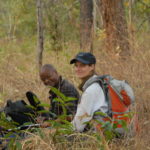 A white woman and a Black man sitting among grasses, shrubs and trees during fieldwork