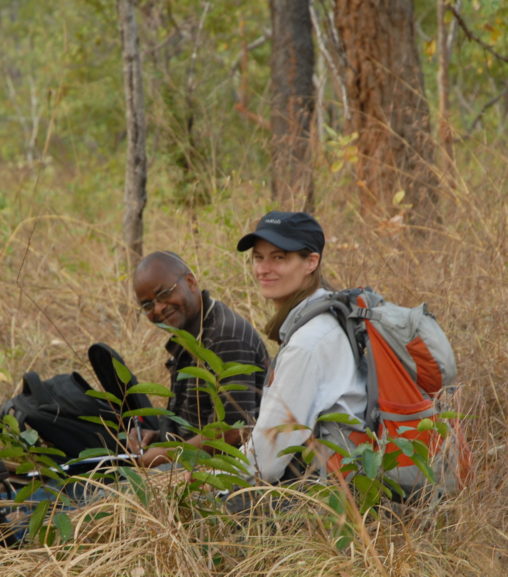 A white woman and a Black man sitting among grasses, shrubs and trees during fieldwork