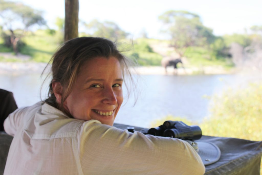 A woman in a white shirt smiling back at the camera. She is sitting in a hide from which an elephant can be seen in the distance