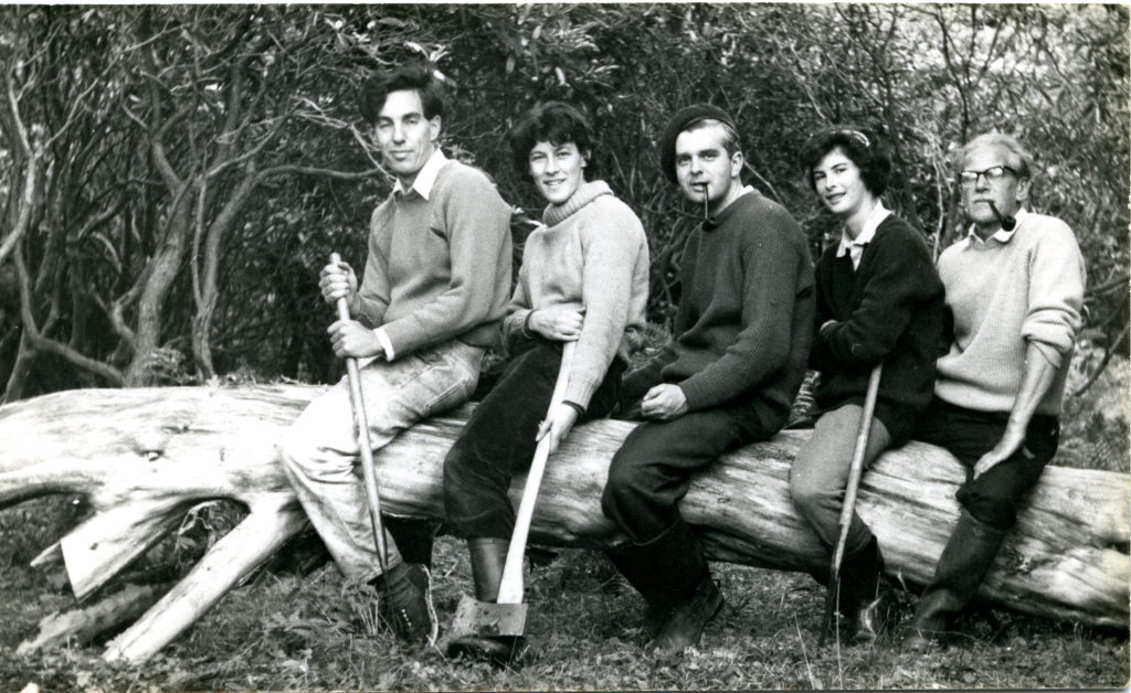 A black-and-white photograph of three men and two women sitting astride a fallen tree trunk. Some of them hold gardening tools. The two Andrews are smoking pipes.
