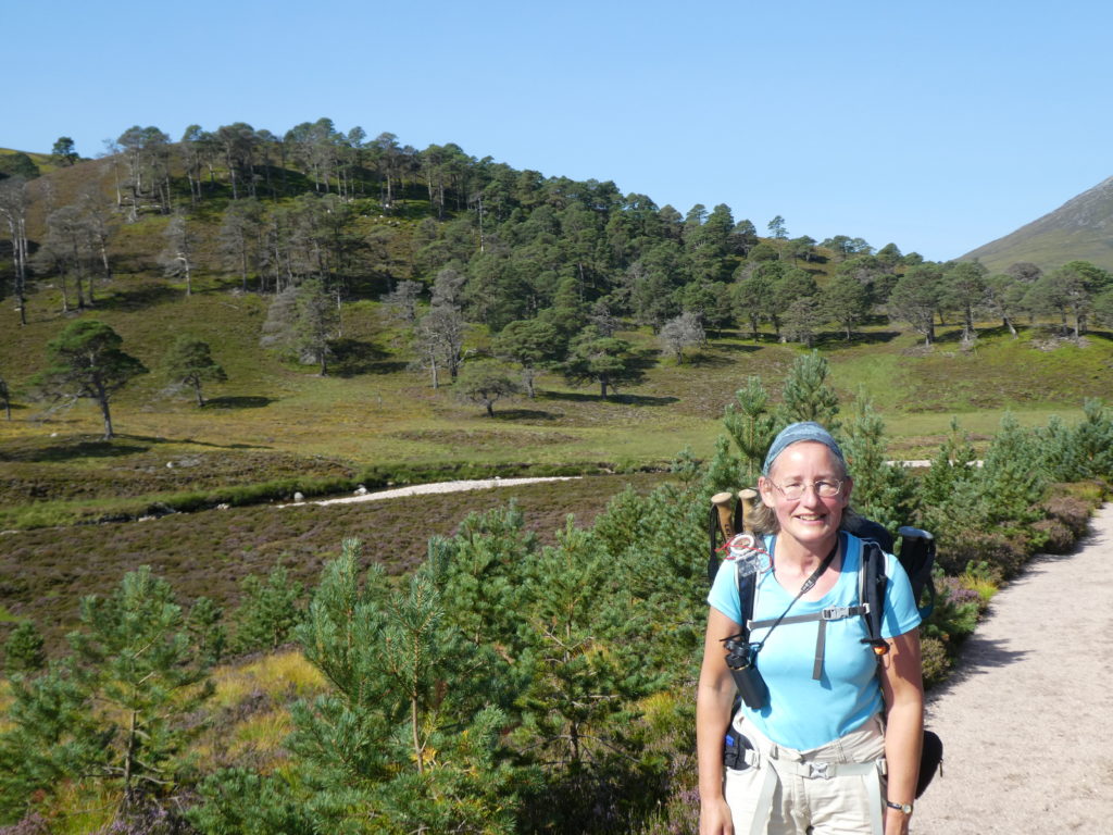A lady in a blue t-shirt wearing glasses, a backpack and carrying binoculars stands in front of a wooded hillside with a blue sky beyond.