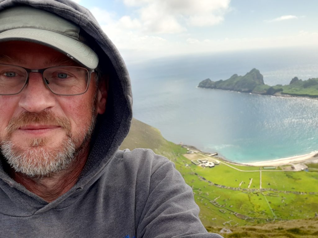 A white man with a short beard wearing a green cap and grey hoodie pulled over it, stands in front of a beautiful bay with green grass, white sand and blue sea