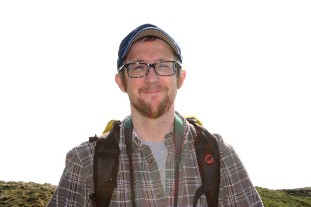 A white man with a beard and glasses, wearing a blue cap and checked grey shirt, carrying a backpack and smiling at the camera