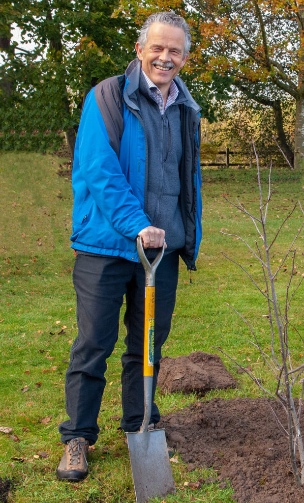 A smiling man in a blue jacket with a spade and a just-planted sapling.