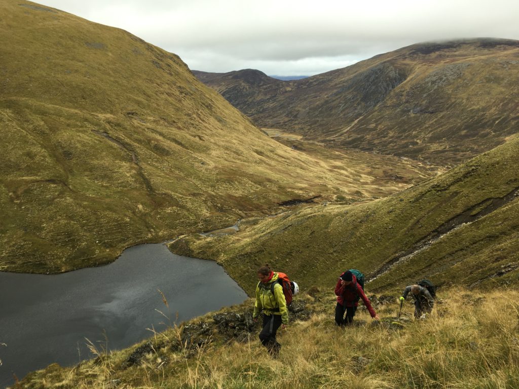 Three people walking up a steep grassy slope in the Highlands, with a small loch in the valley below. Aline is in the front, in a yellow jacket with a backpack on