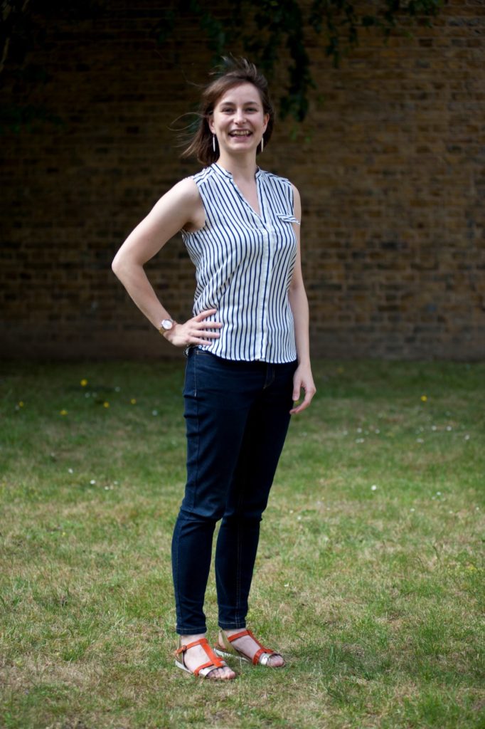 A smiling white woman with brown hair, wearing jeans and a sleeves white-and-navy striped shirt, with her right hand on her hip