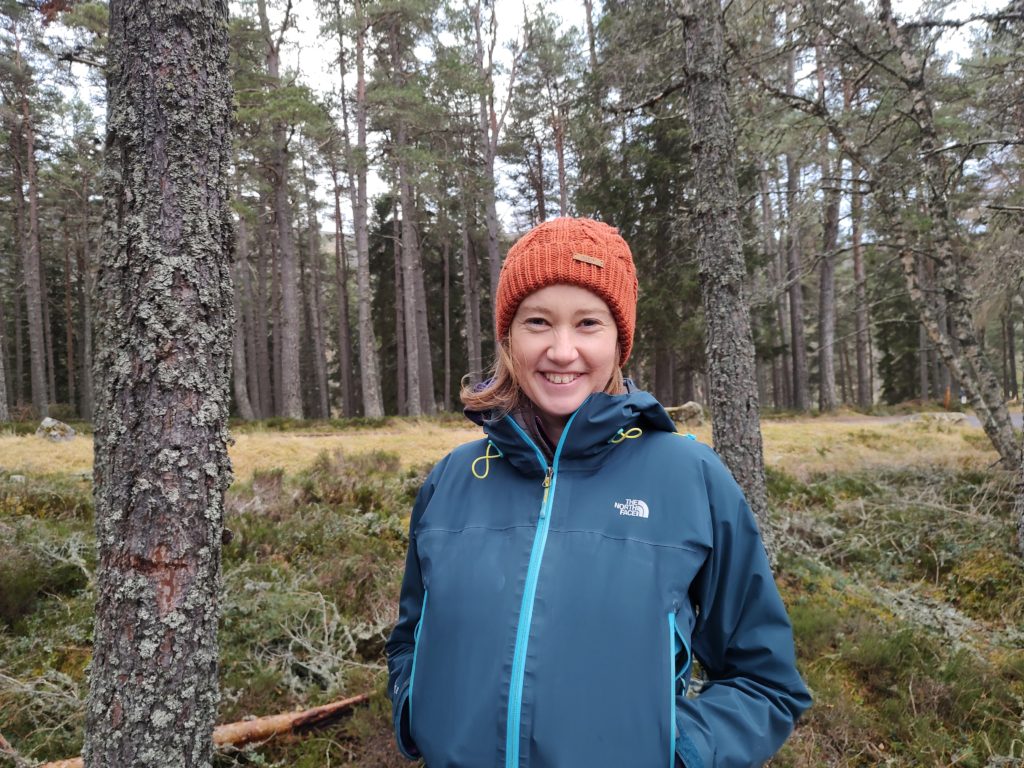 A smiling lady wearing an orange woolly hat and blue jacket stands in a woodland with lichen-covered trunks all around