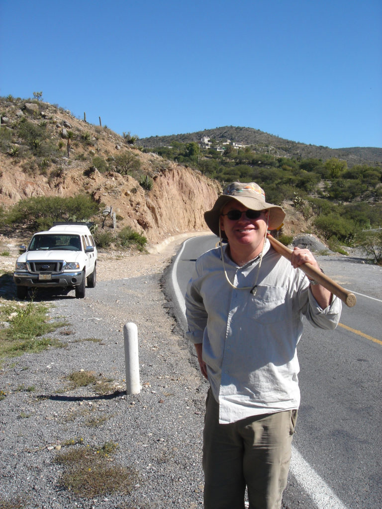 A white man wearing fieldwork clothes, sunglasses and a floppy hat, carrying a tool over his shoulder, stands by a road with a parked 4WD in a dry, mountain landscape