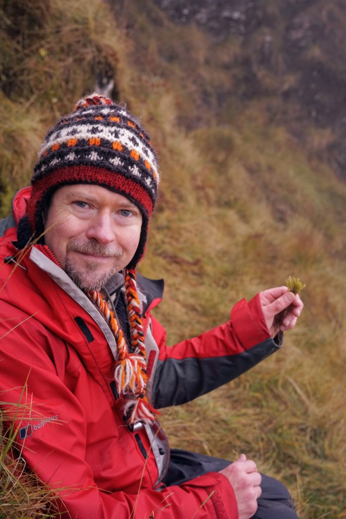 A white man sitting in a mountain environment, wearing a red jacket and multicoloured woolly hat, smiling at the camera and holding a bryophyte in his hand