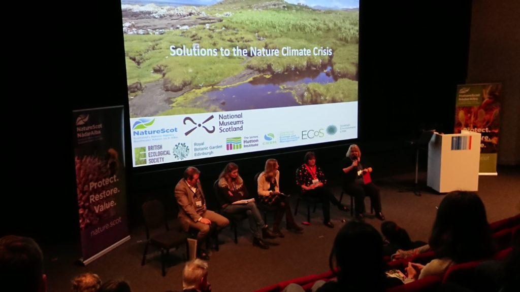 A panel of five people sit on chairs in front of a screen saying 'Solutions to the nature climate crisis' over a Scottish landscape image