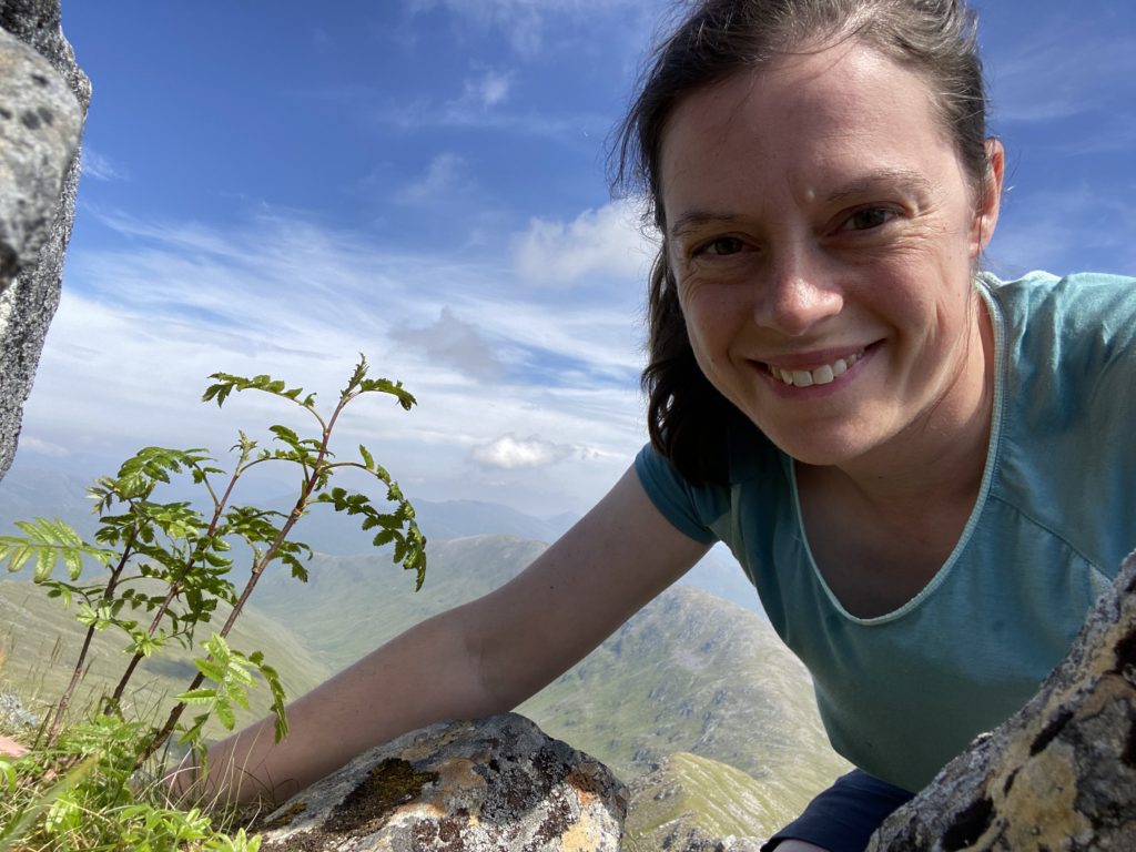 A woman smiles from a mountain with a blue sky and wispy clouds behind and a young rowan sapling next to her