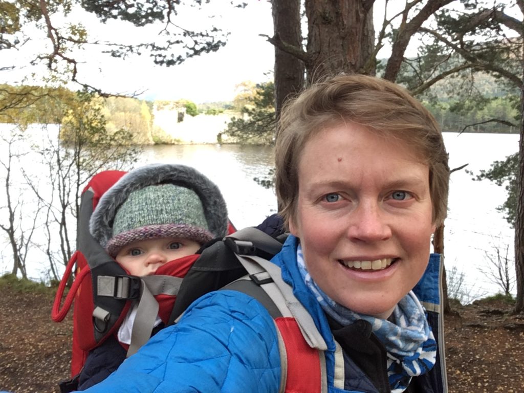 A head-and-shoulders shot of a smiling woman in front of a loch with an island in the middle of it. She has a baby-carrier on her back with a baby in a woolly hat peeking out of it.
