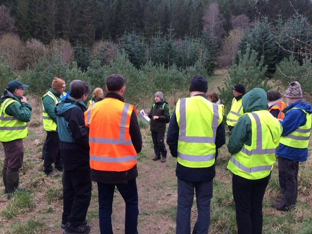 A group of people in hi-vis jackets with Joan at the centre, talking to them, in a landscape with young trees