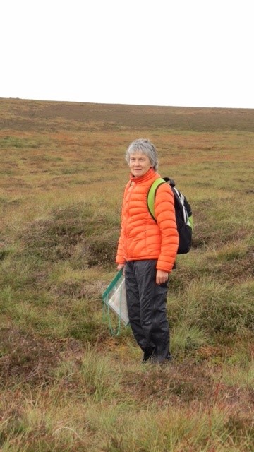 A lady in a bright orange jacket, carrying a backpack and map, stands in a moorland landscape