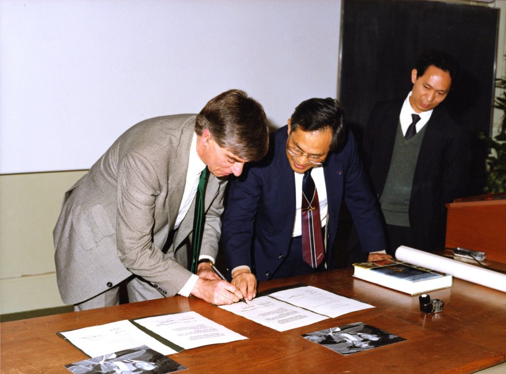 Two men in suits and ties leaning over a document on a table, the one on left holds a pen. Another man looks on from behind