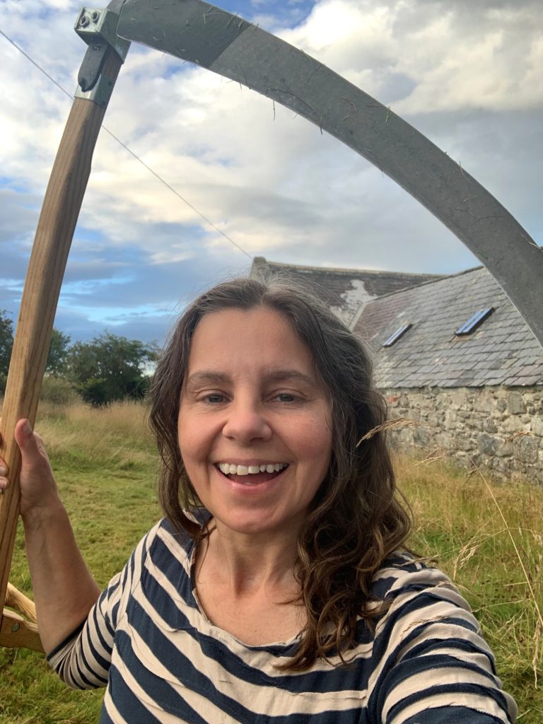 A lady with long, loose brown hair, wearing a striped top, wielding a scythe in a grassland in front of a stone building