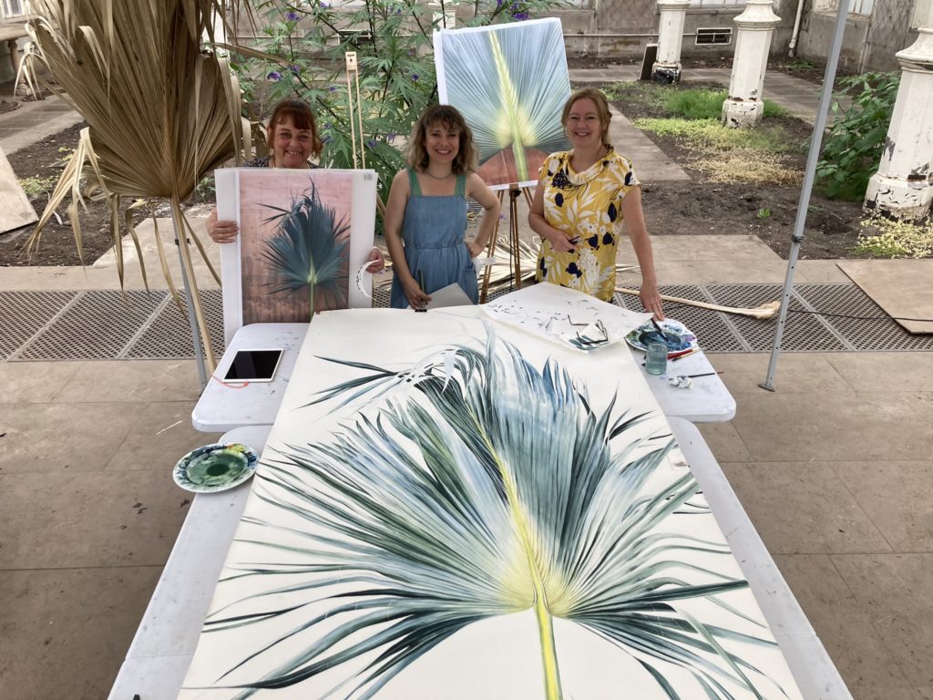 A large painting of a palm frond, in front of 3 female artists