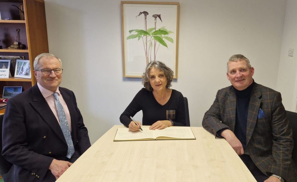 Three people sit around the head of a table: from left, Simon Milne, in a suit and tie wearing glasses, Sandra Díaz, wearing a black dress with tartan trim, signing a guest book on the table in front of her, Dominic Fry in a jacket and black sweater.