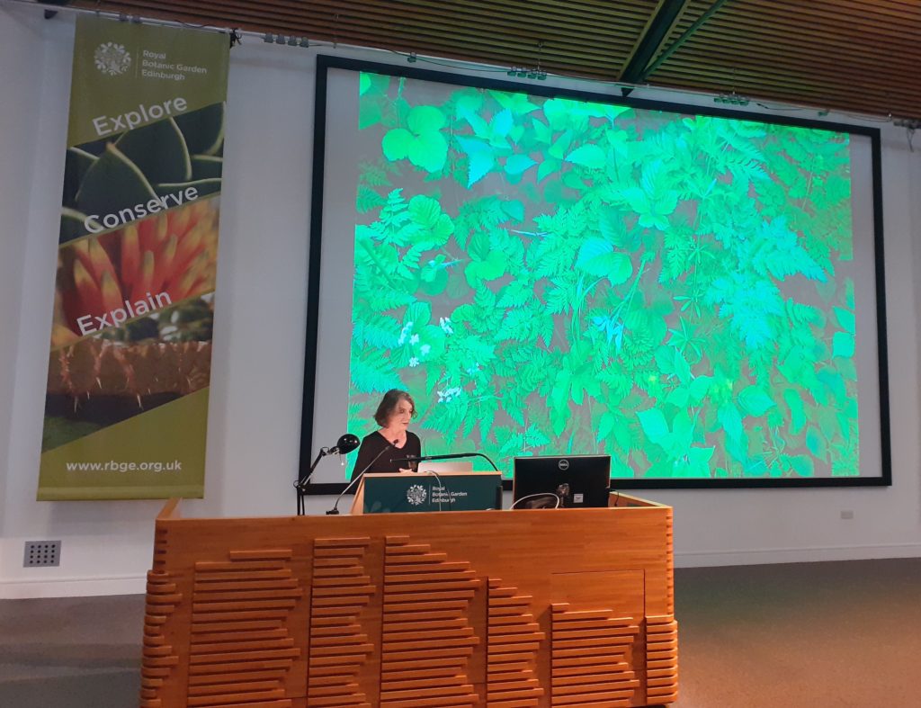 Sandra Díaz standing at a lectern in front of a slide showing a community of herbaceous plants