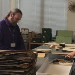 Chris Knowles (left) and Robyn Drinkwater (right) recurating RBGE Magnoliaceae herbarium specimens on a sorting bench. There are folders stacked at the front of the bench and herbarium folders and other stationary supplied spread across the bench(table) surface.
