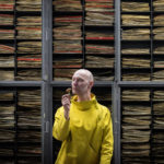 A bald white man wearing a bright yellow smock stands in front of a series of grey cabinets, which are open to reveal multiple herbarium folders stacked inside. He holds in his right hand a dried mushroom specimen, which he has raised up to his nose as if to smell it.
