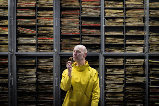 A bald white man wearing a bright yellow smock stands in front of a series of grey cabinets, which are open to reveal multiple herbarium folders stacked inside. He holds in his right hand a dried mushroom specimen, which he has raised up to his nose as if to smell it.