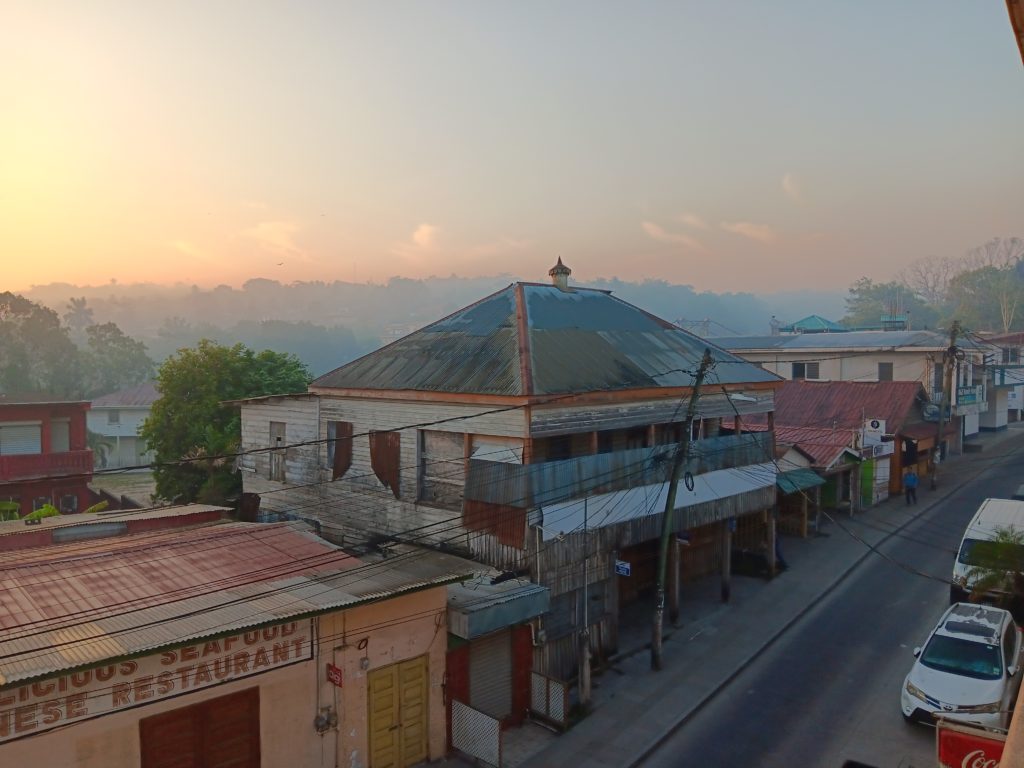 Dawn over a tropical town with low roofs of rusty corrugated iron and a quiet street in the foreground. A tree-covered hill in the background.