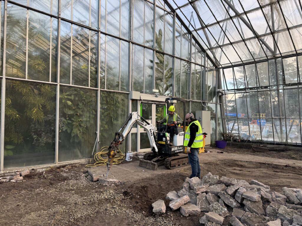 Two men standing, one a digger breaking down a path in a glasshouse