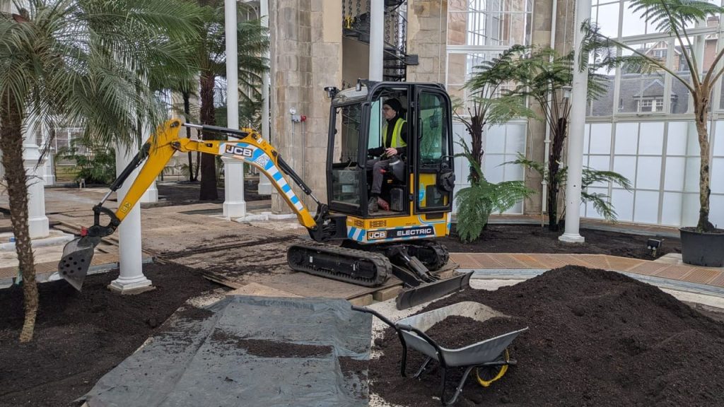 A man operates a digger, moving soil from one area to another inside the Palm House.