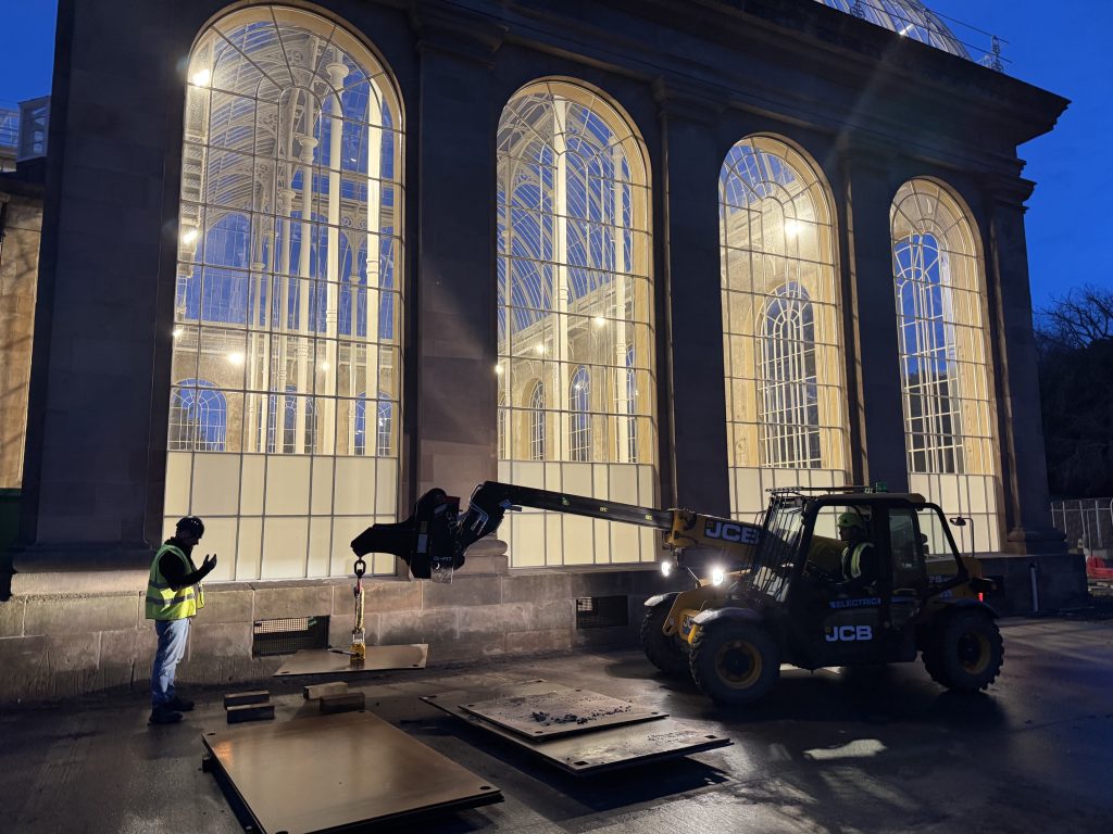 Dark morning the Palm Houses in the background light up, in the foreground two horticulturists move steel plates.